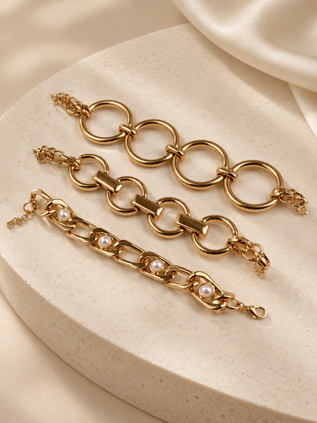 Três pulseiras douradas em tamanhos diferentes, com elos circulares e detalhes em pérolas